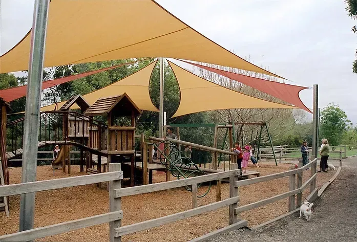 Custom-designed yellow and orange shade sails installed over children’s playground at Berwick Botanical Garden, supported by tall steel posts and covering timber play structures including slides and climbing frames, providing full UV protection in a landscaped public setting