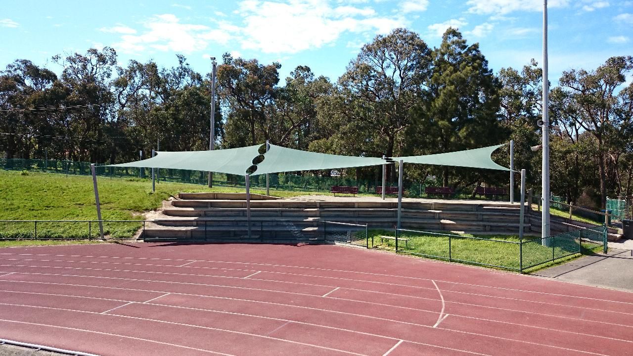 Shade sail installation at Berwick High School athletics track, Manuka Rd, Berwick VIC 3806—featuring a large triangular canopy tensioned across tall steel posts, offering sun protection over a grassed viewing area beside the track, surrounded by native trees, fencing, and open sky, enhancing outdoor sports facilities for students