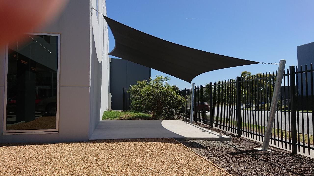 Industrial shade sail installation in Pakenham—custom black canopy tensioned between steel posts and a factory façade, creating a sheltered staff seating area in a new commercial development with red benches, stone seating, and enhancing the surrounding space and business 