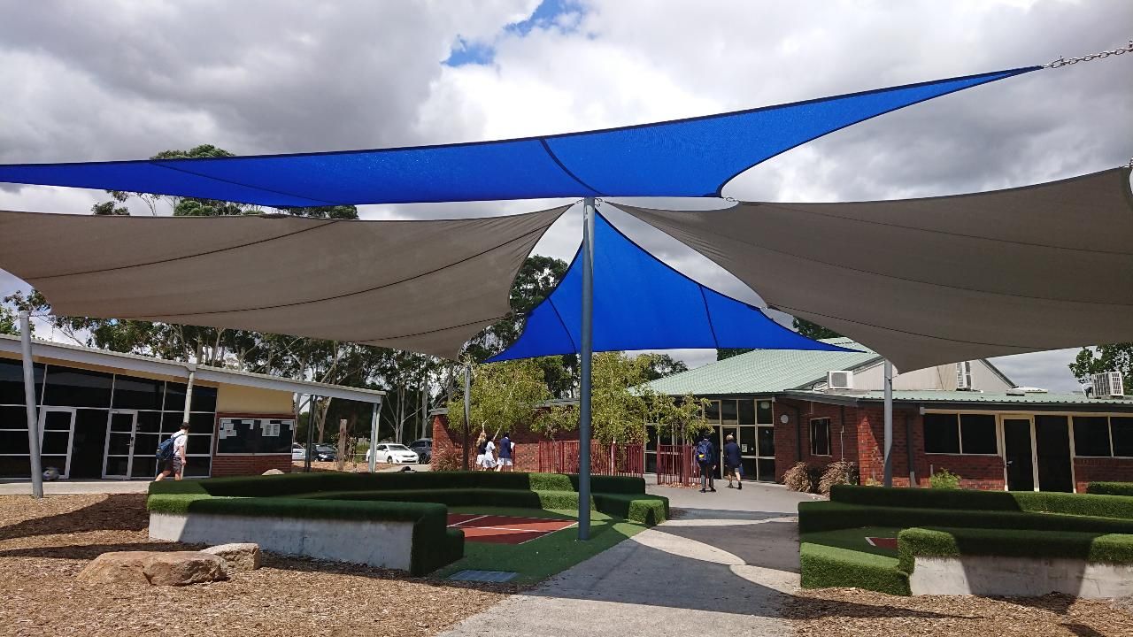 Multiple beige and blue shade sails installed over the amphitheatre seating area in the open play spaces at Beaconhills College Pakenham, supported by metal posts and providing UV protection over artificial turf and concrete benches with school buildings in the background