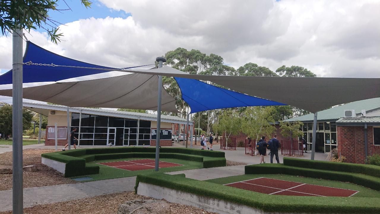 Custom shade sail canopy system covering outdoor amphitheatre at Beacon Hills College in Pakenham, featuring tensioned fabric in blue and beige tones over landscaped green play space with surrounding educational facilities.