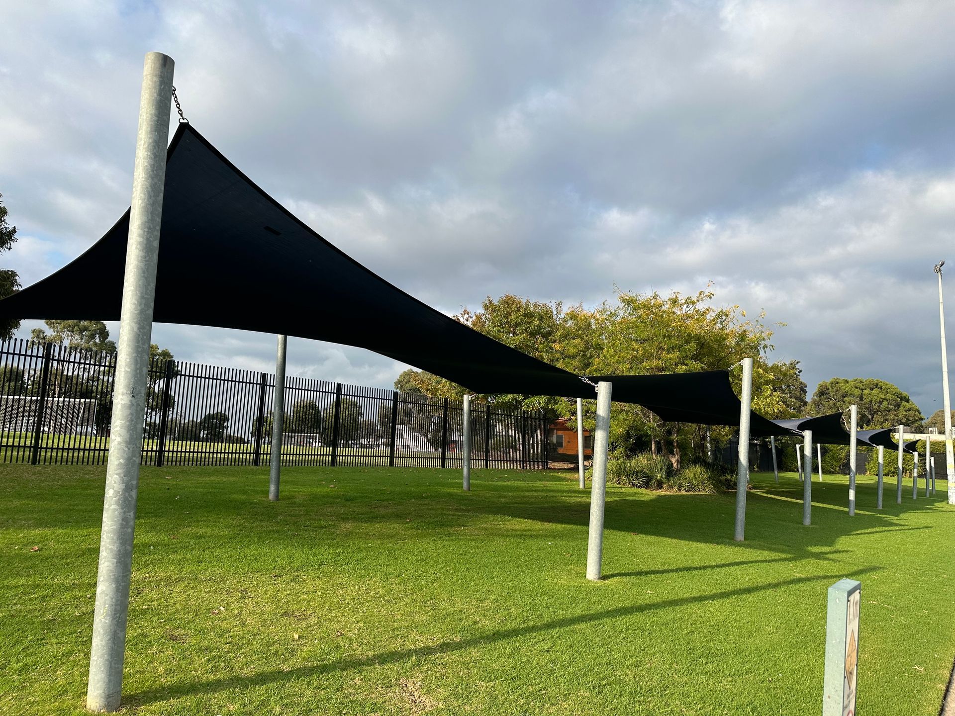 Commercial shade sail installation at Doveton Pool in the Park, Kidds Rd & Tristania St, 64 Tristania St, Doveton VIC 3177—featuring multiple linked hyperbolic canopies tensioned across tall steel posts, creating expansive sun coverage over a grassy area beside fencing and trees