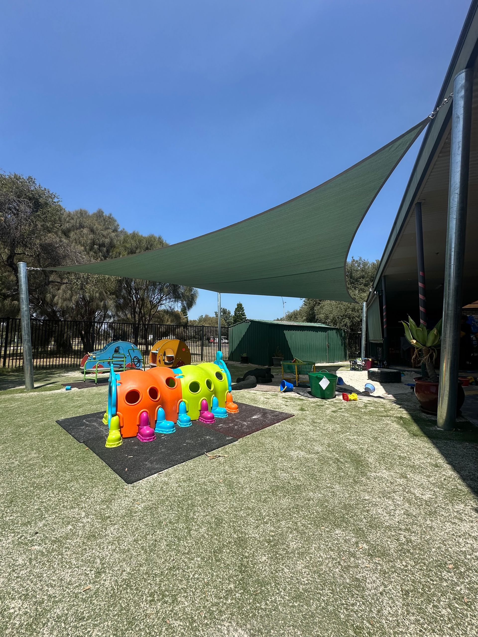 Shade sail installation at Port Melbourne Kindergarten, Melbourne 3150, featuring a durable green canopy providing sun protection over a vibrant playground with colourful equipment, artificial grass, and surrounding trees. Custom-built by a licensed shade sail installer in Melbourne