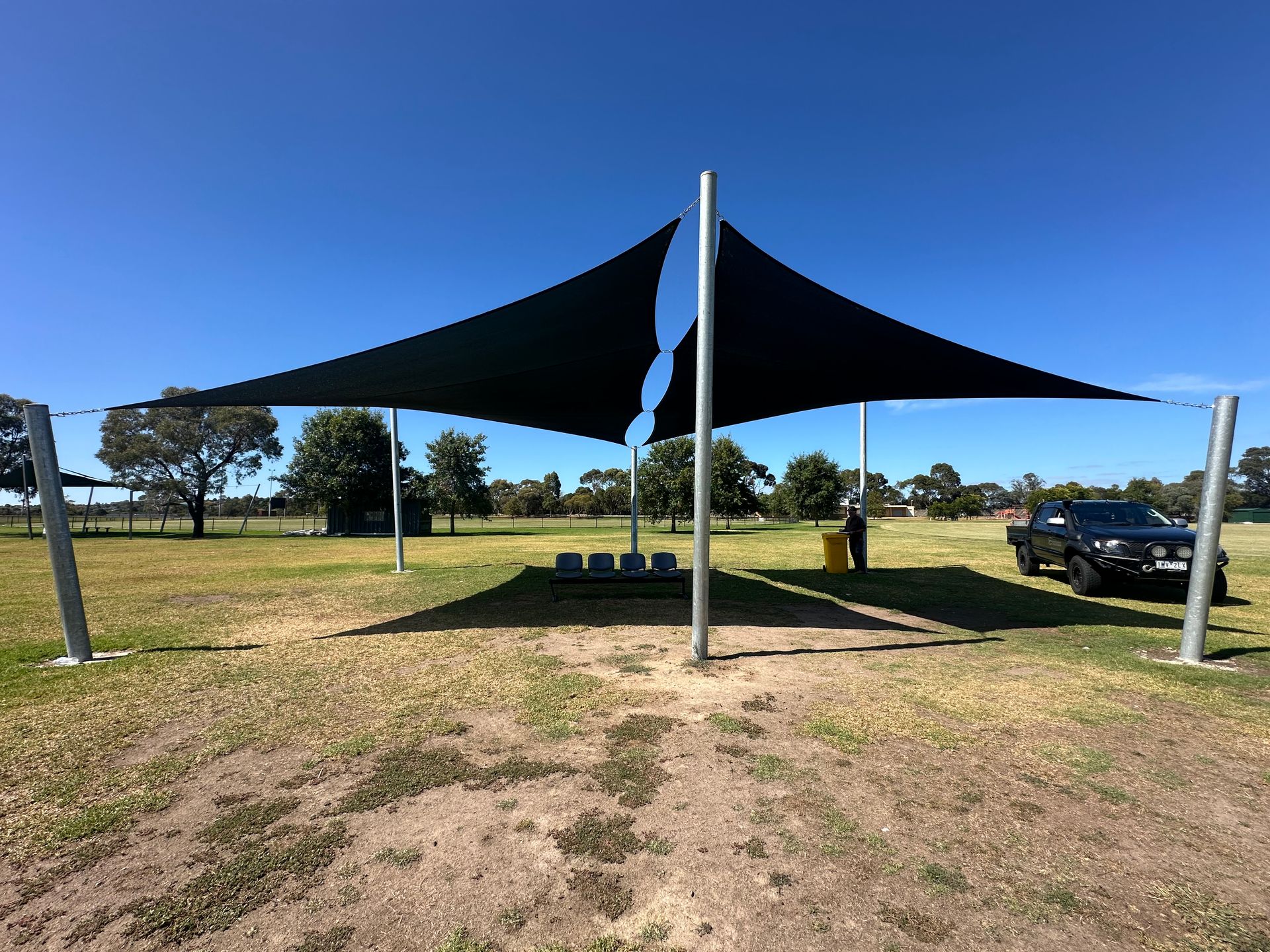 Custom commercial shade sail installation at Sydney Pargeter Reserve Playground, Sydney Pargeter Recreation Area, Kennington Park Dr, Endeavour Hills VIC 3802—featuring two joined square hyperbolic twist canopies over each football oval, tensioned between steel posts to create dynamic architectural shade for public seating areas, with fencing, trees, and recreational facilities in the background