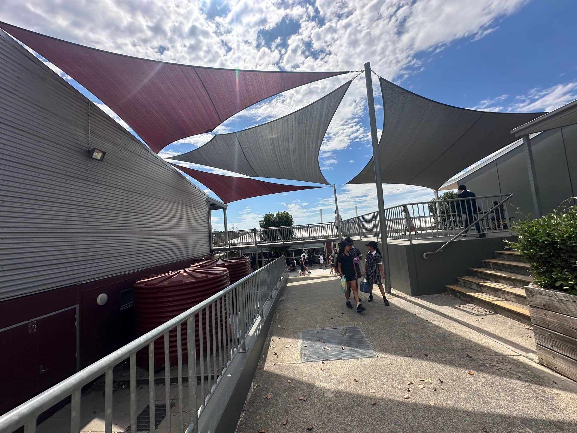 Custom shade sail canopy system covering outdoor amphitheatre at Beaconhills College in Berwick, featuring tensioned fabric in maroon and beige tones over landscaped green play space with surrounding educational facilities.