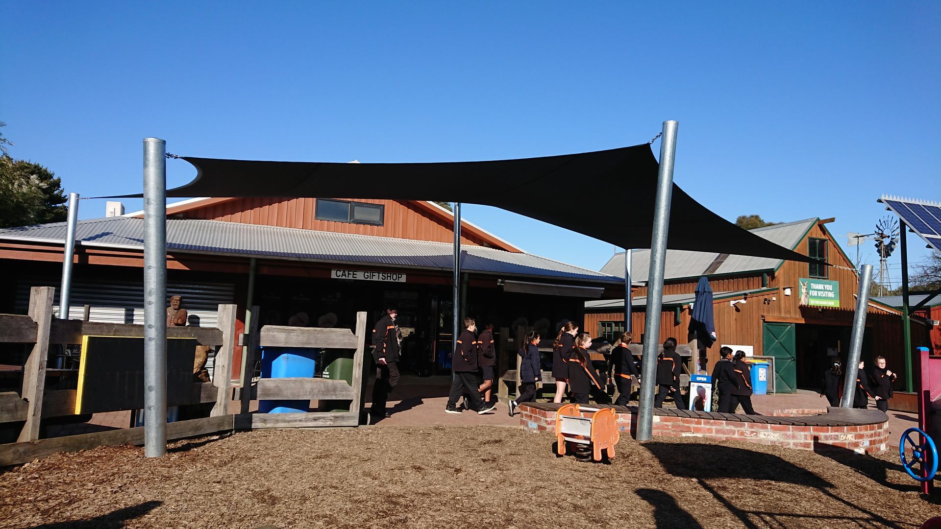 Shade sail installation at Myuna Farm, 182 Kidds Rd, Doveton VIC 3177—featuring a large beige canopy supported by steel posts, shading the cafe’s outdoor seating area adjacent to a family-friendly playground with green spaces and rustic farm surroundings