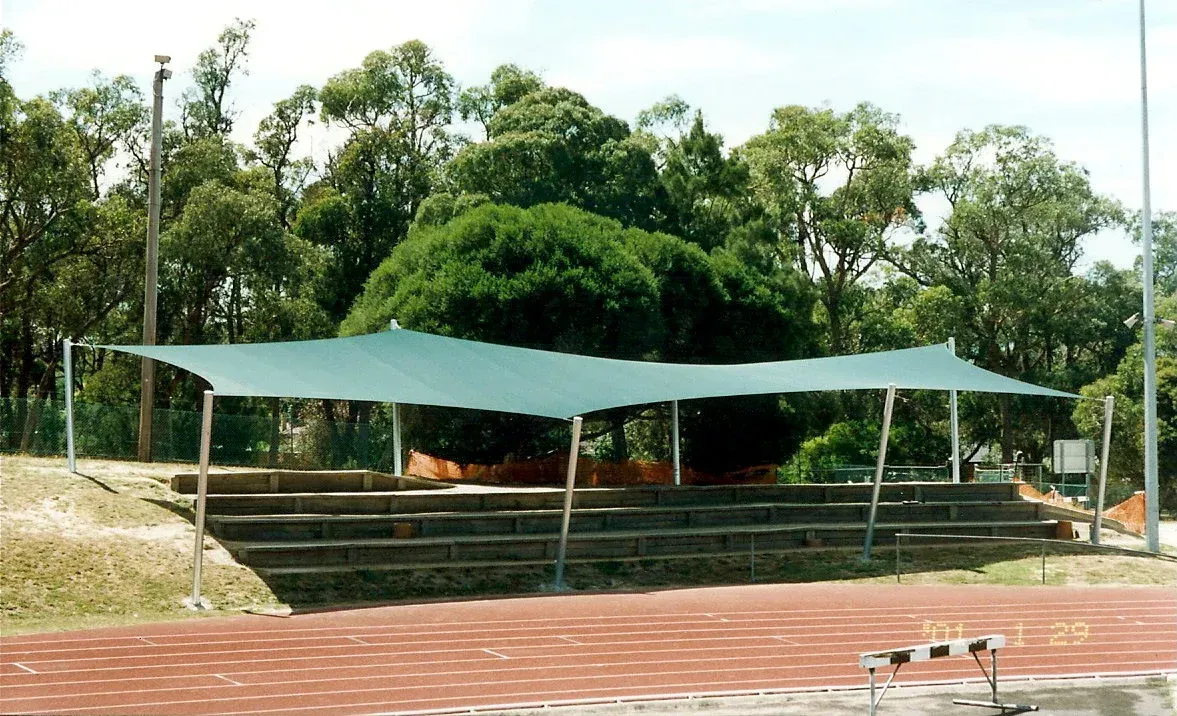 Shade sail installation at Berwick High School athletics track, Manuka Rd, Berwick VIC 3806—featuring a large triangular canopy tensioned across tall steel posts, offering sun protection over a grassed viewing area beside the track, surrounded by native trees, fencing, and open sky, enhancing outdoor sports facilities for students