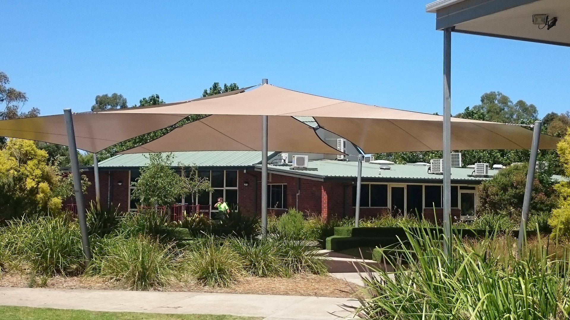 Custom shade sail canopy system covering outdoor amphitheatre at Beaconhills College in Pakenham, featuring tensioned fabric in blue and beige tones over landscaped green play space with surrounding educational facilities.