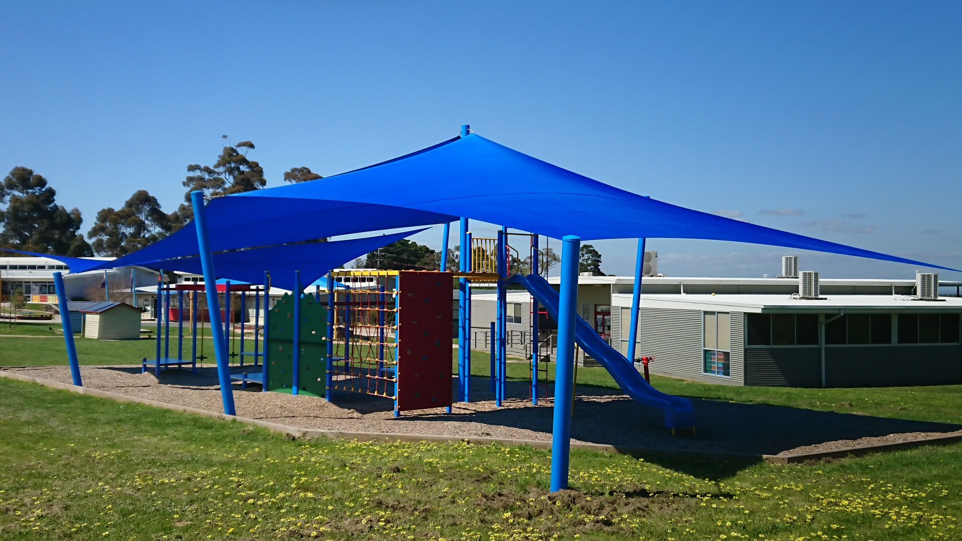 Large blue commercial shade sail installed over a spacious school playground at St Brigid’s Catholic Primary School, 25 Bayview Rd, Officer VIC 3809. The shade structure is anchored by tall blue posts and covers a variety of play equipment including climbing frames, a slide, and rope nets. The installation provides effective UV protection while enhancing outdoor comfort and safety, professionally completed by a licensed Melbourne shade sail specialist