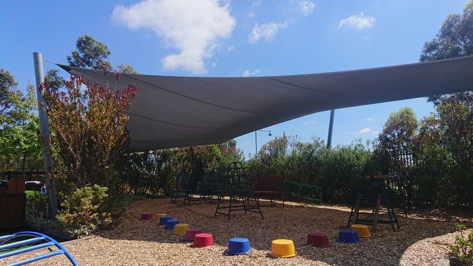 Custom-installed dark shade sail at a kindergarten in Clyde North, Melbourne VIC 3978, covering a natural play area with climbing equipment and colourful stepping stones. Surrounded by native greenery and trees, the sail provides effective UV protection while enhancing the outdoor learning environment. Professionally designed by a licensed shade sail installer in Melbourne