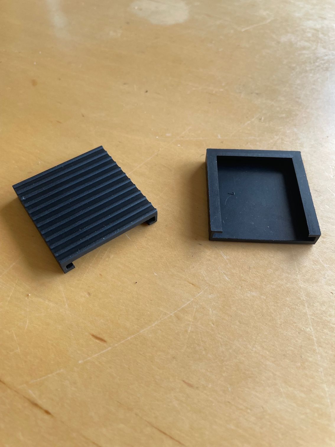 Two black square rubber furniture feet on a light wood surface. One foot has ridges.