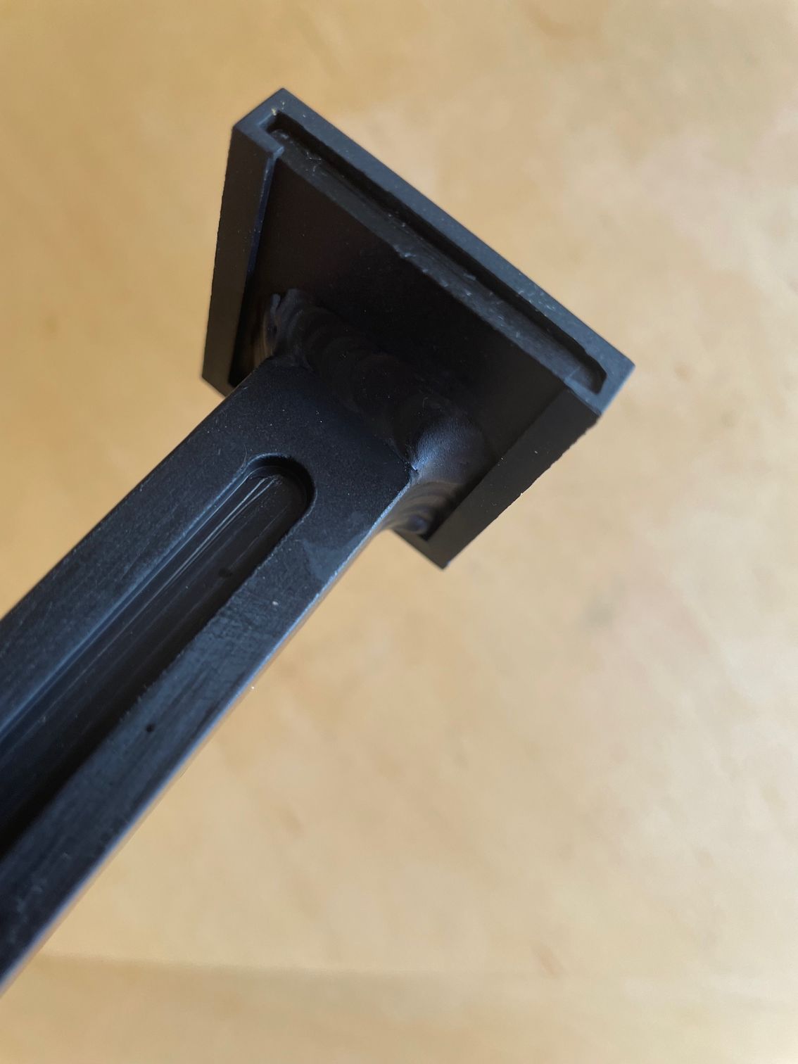 Black, T-shaped 3D-printed part with a rectangular top and a long, grooved stem, resting on a wooden surface.