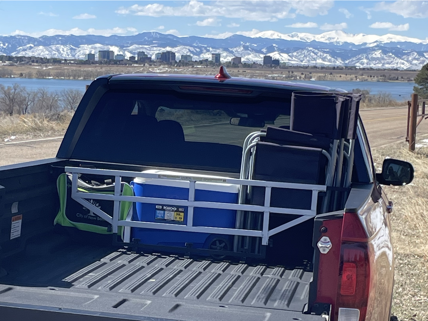 Truck bed with camping gear: cooler, chairs, and bag. Scenic view with mountains and water in the background.