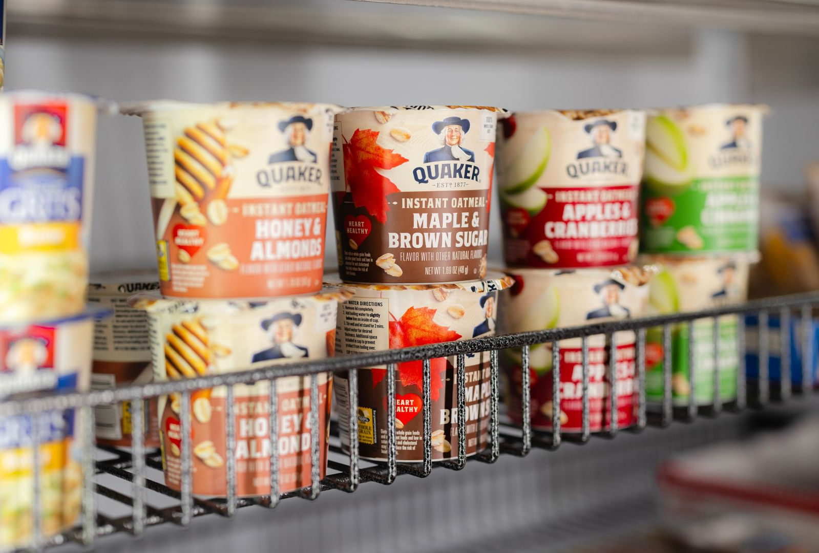 A store shelf displays rows of Quaker Instant Oatmeal cups in flavors like Honey & Almonds, Maple & Brown Sugar, and Apple.