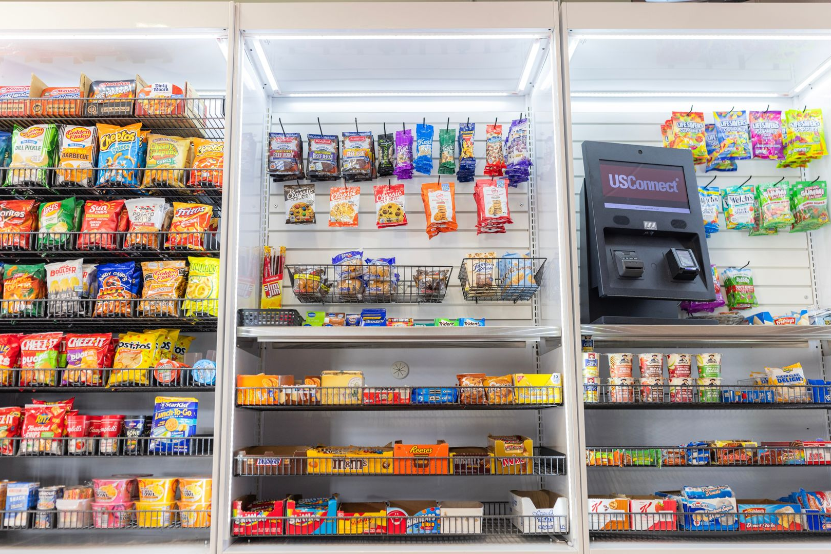 A well-stocked convenience store snack aisle with shelving displays of various packaged chips, candy, and a checkout kiosk.
