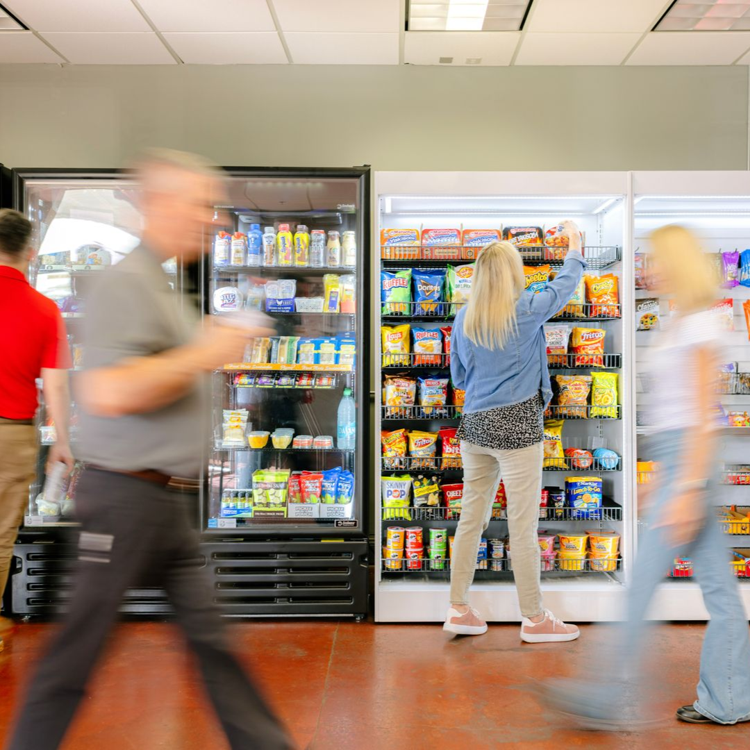 Three people walk past or browse in front of two glass-fronted vending machines filled with snacks in a bright office.