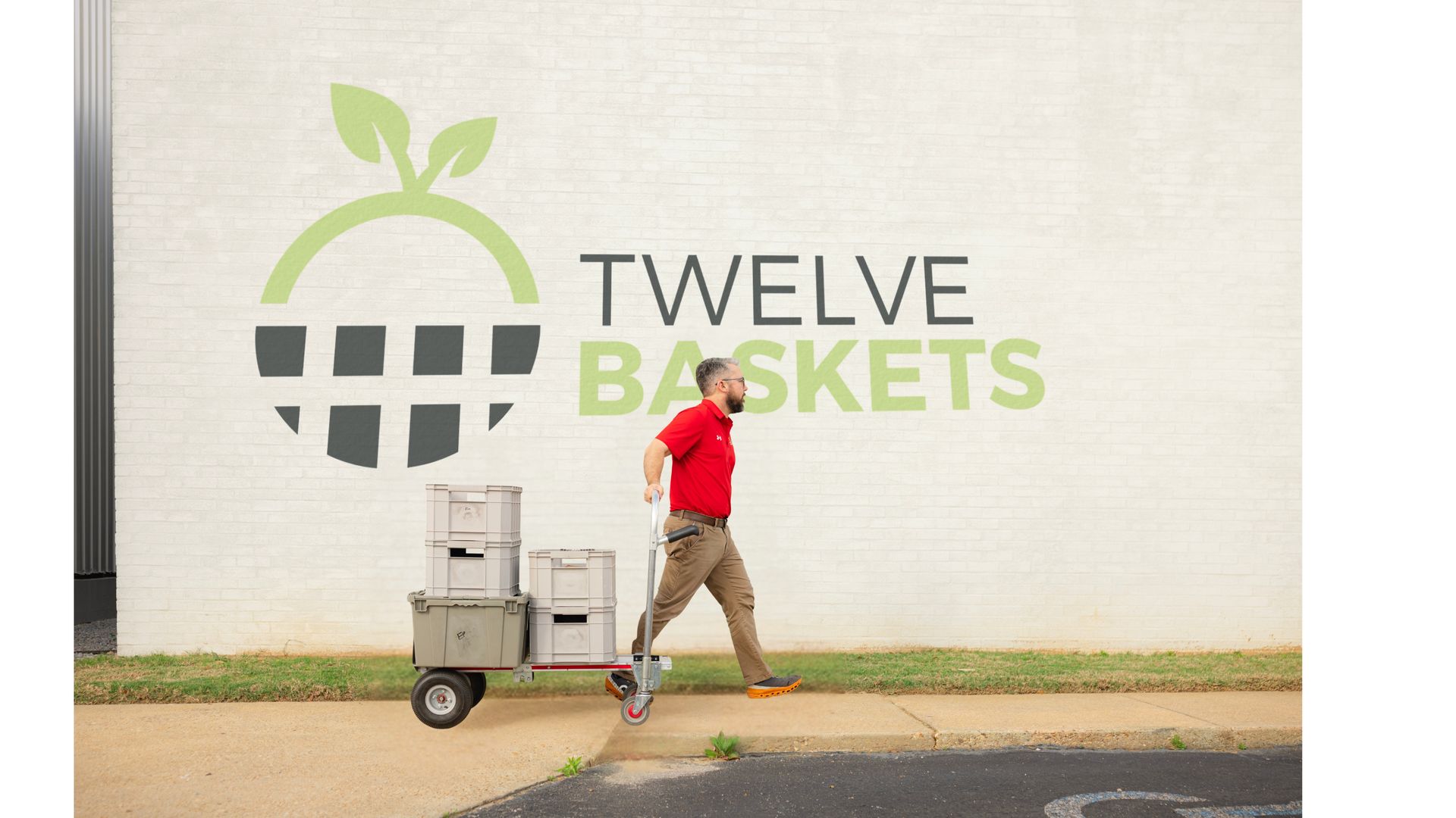 A person in a red shirt pulls a cart with crates along a wall featuring a Twelve Baskets logo.