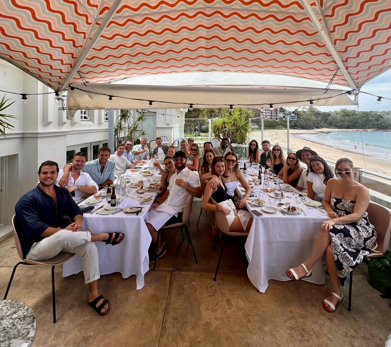 A group of people are sitting at a long table under an umbrella.