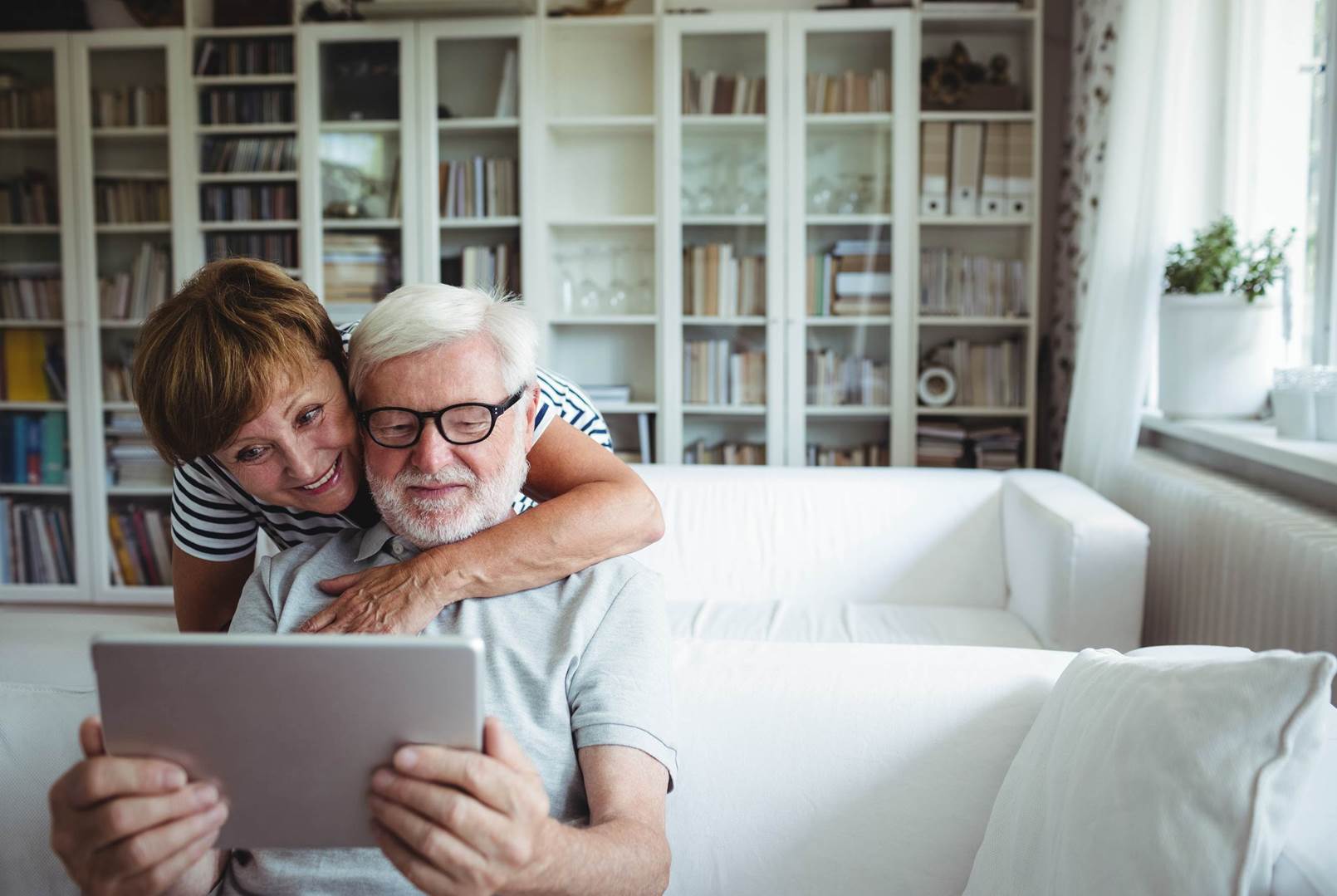 An elderly couple is sitting on a couch using a tablet computer.