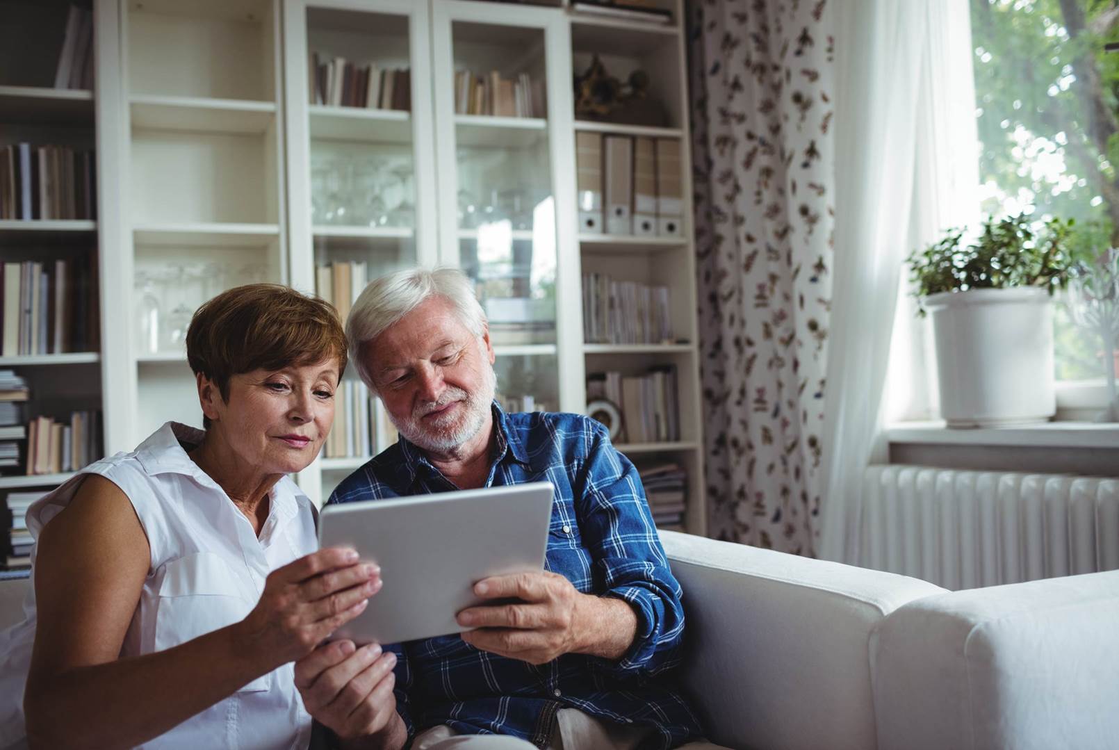 An elderly couple is sitting on a couch looking at a tablet.