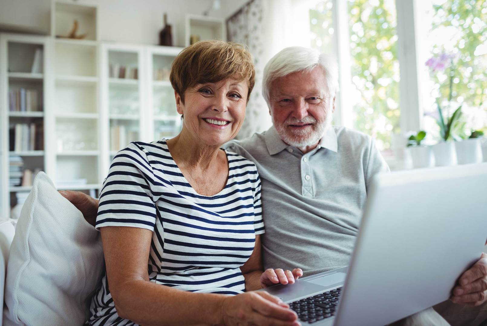 An elderly couple is sitting on a couch using a laptop computer.