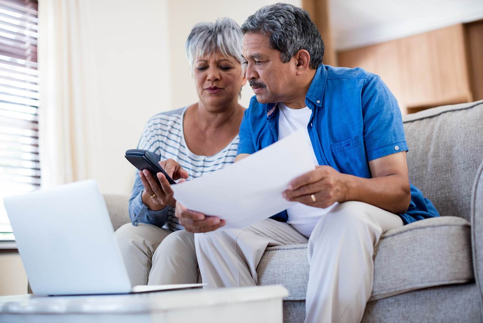 A man and a woman are sitting on a couch looking at a laptop and a cell phone.