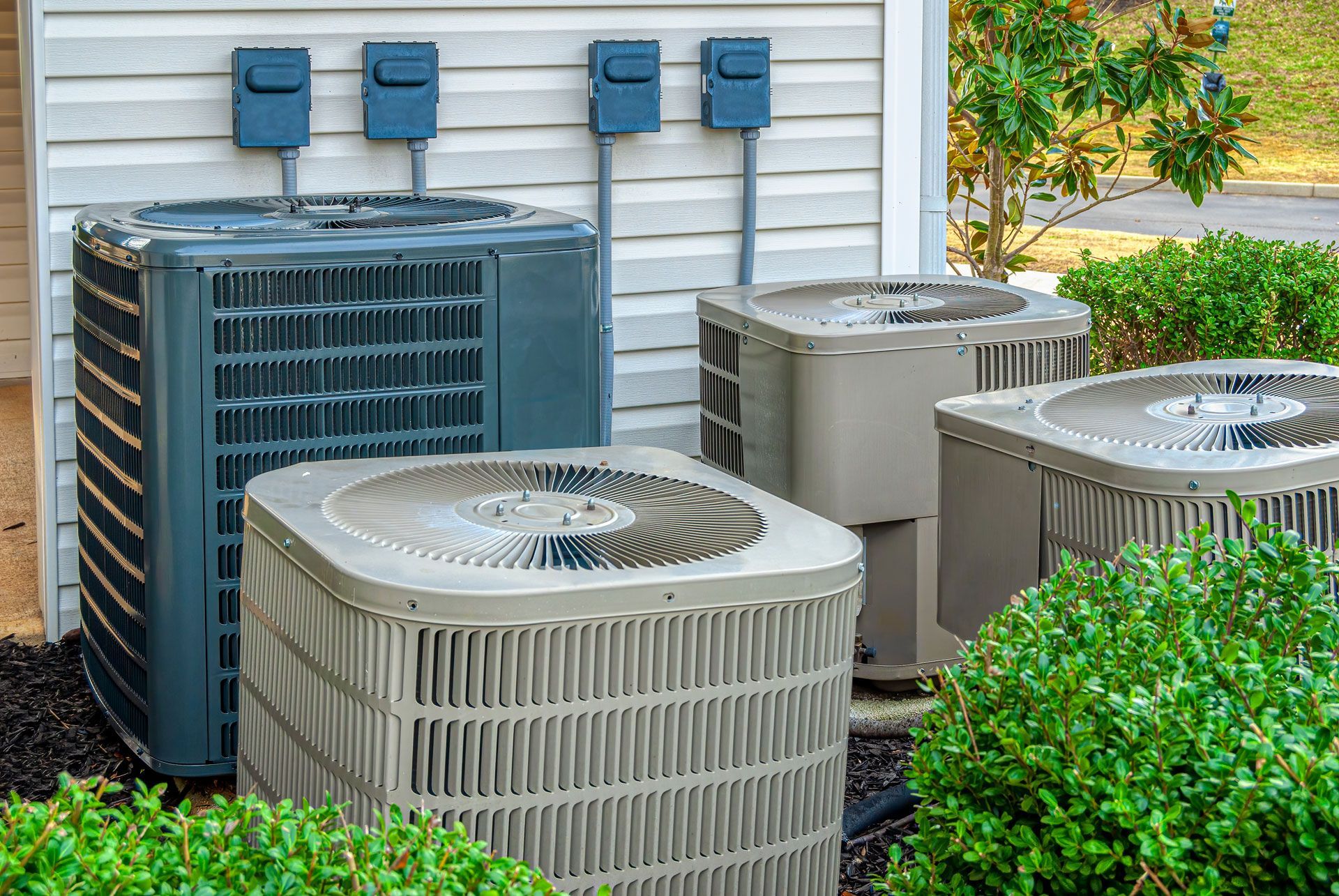 Four air conditioning units outside a building; gray and beige with electrical boxes.