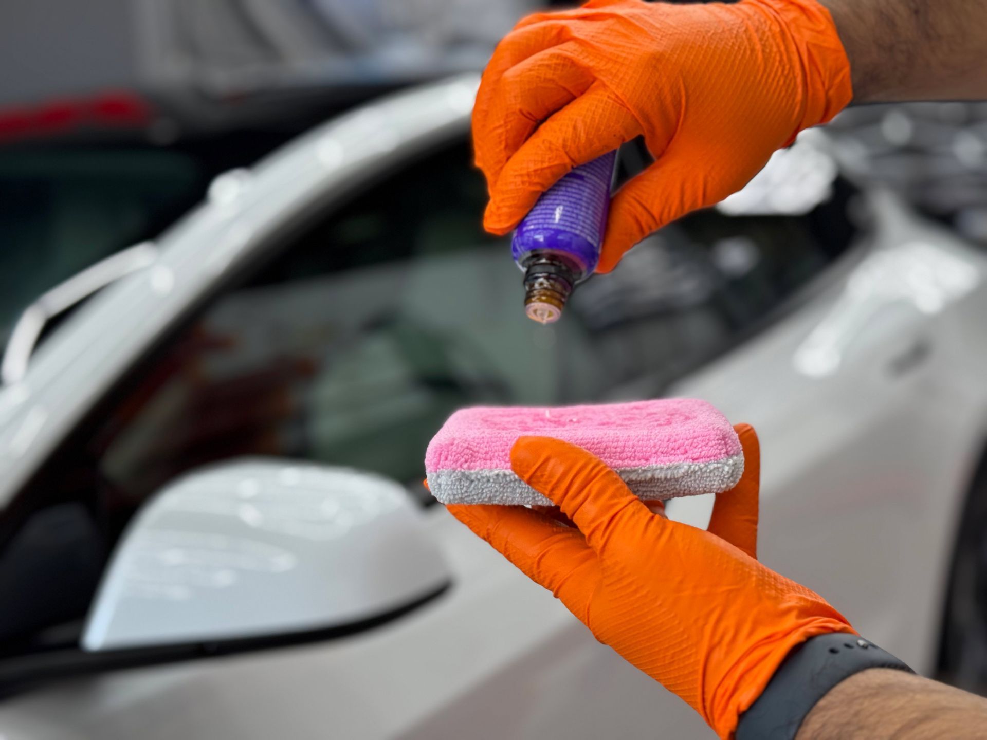 Person wearing orange gloves applying car detailing product to a sponge next to a white car.