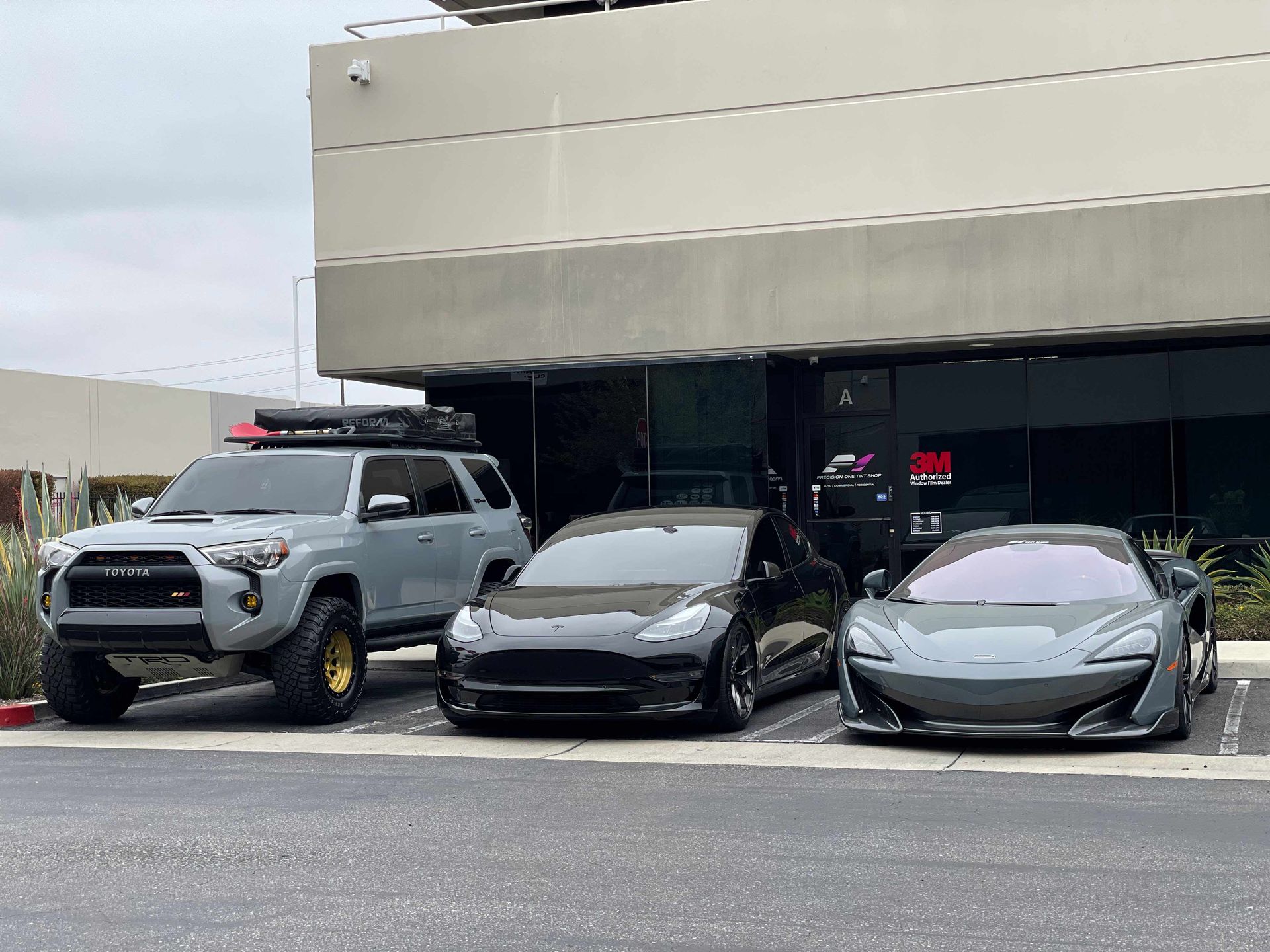 A grey Toyota SUV with roof rack, a black Tesla, and a grey McLaren parked in front of a building.