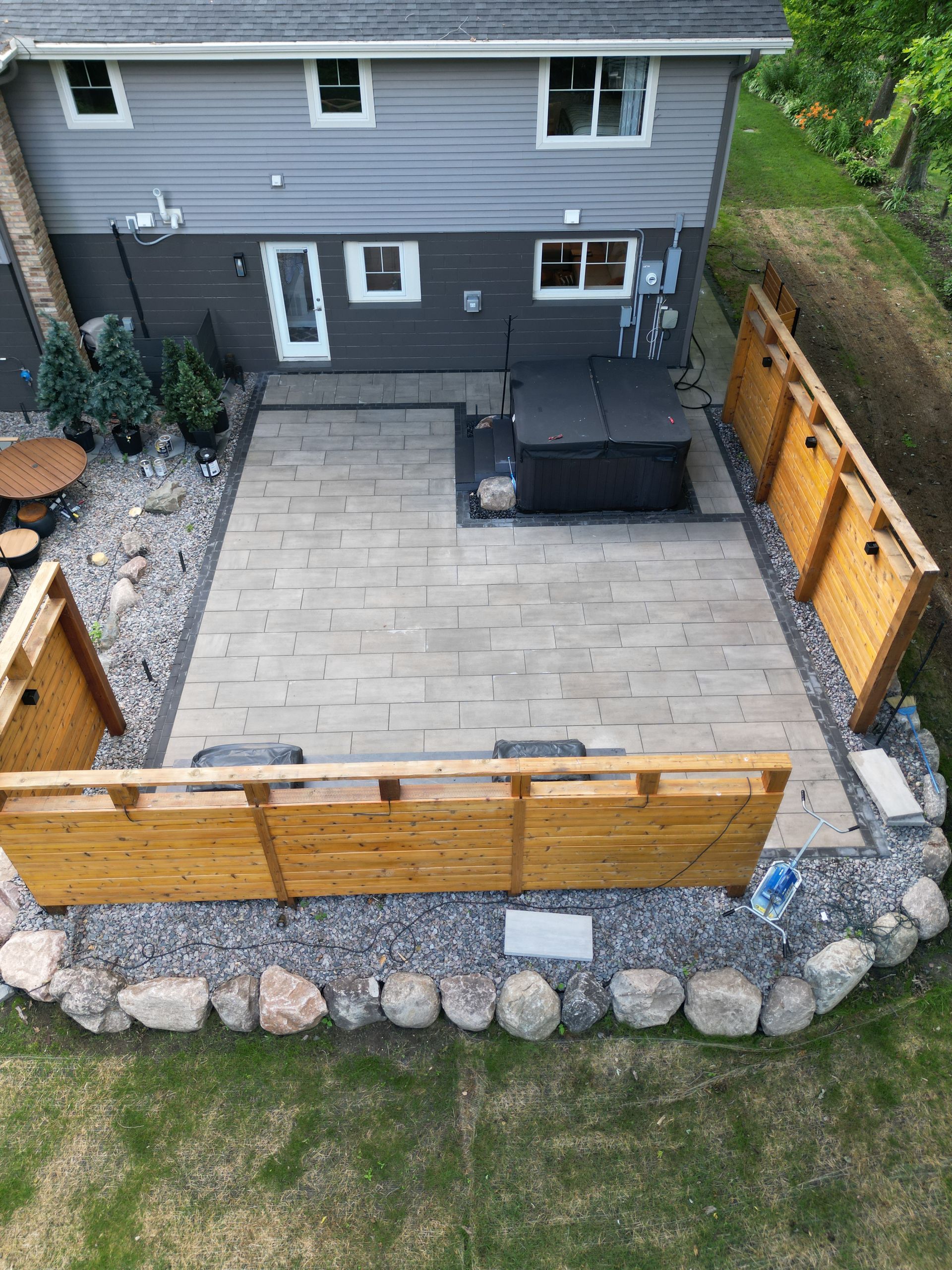 Backyard patio with hot tub, bordered by wood fences and stone. Gray house in background.