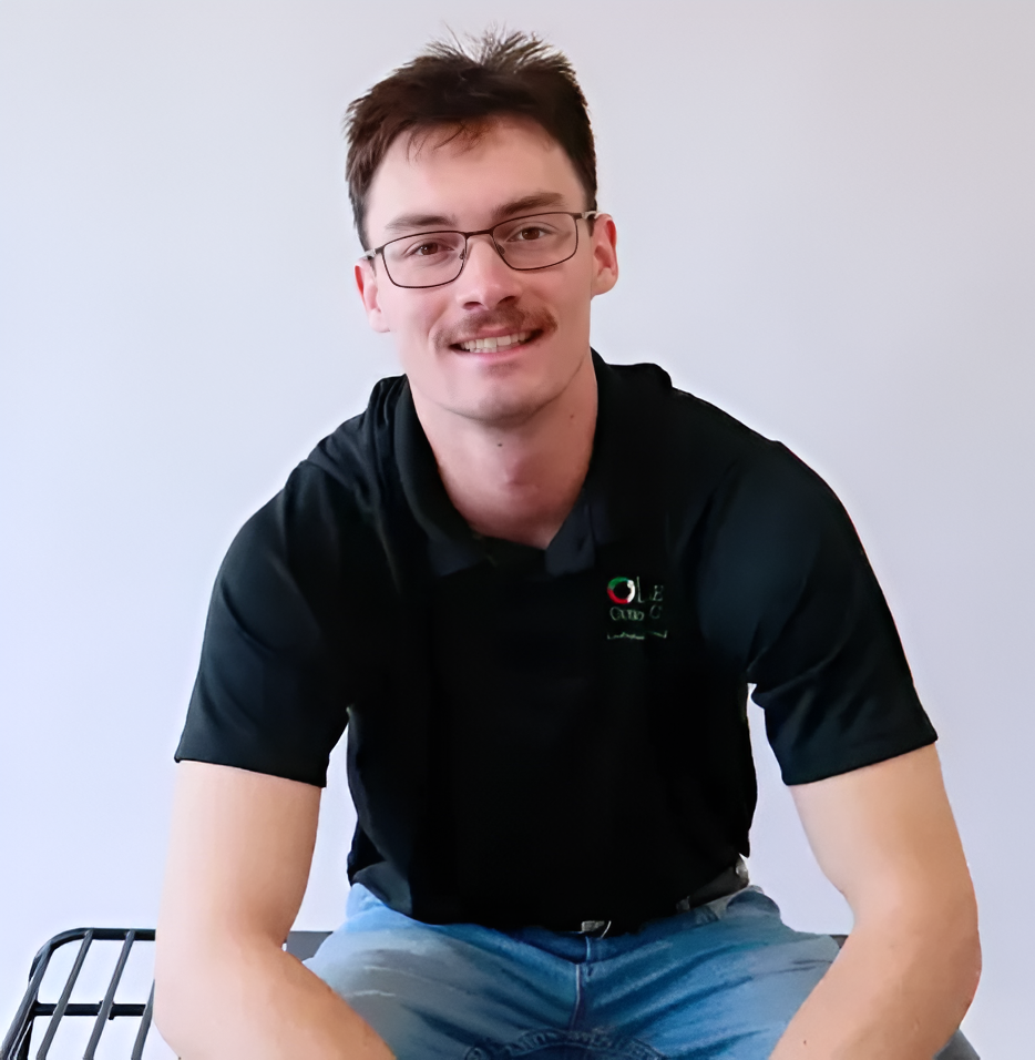 Man with glasses and mustache smiles, wearing a black polo shirt and blue jeans, sitting against a white background.