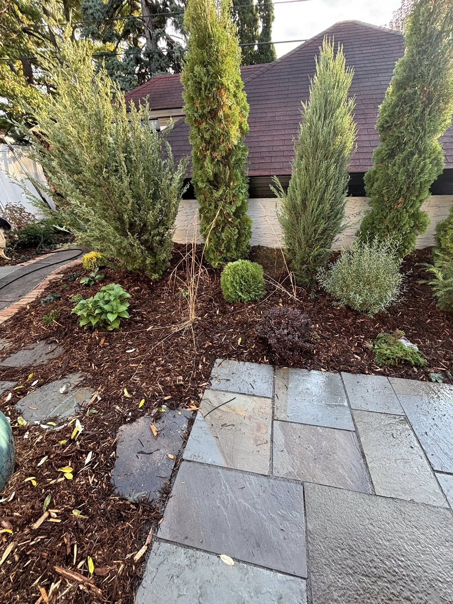 Stone patio leading to a garden bed with varied green shrubs and dark mulch.