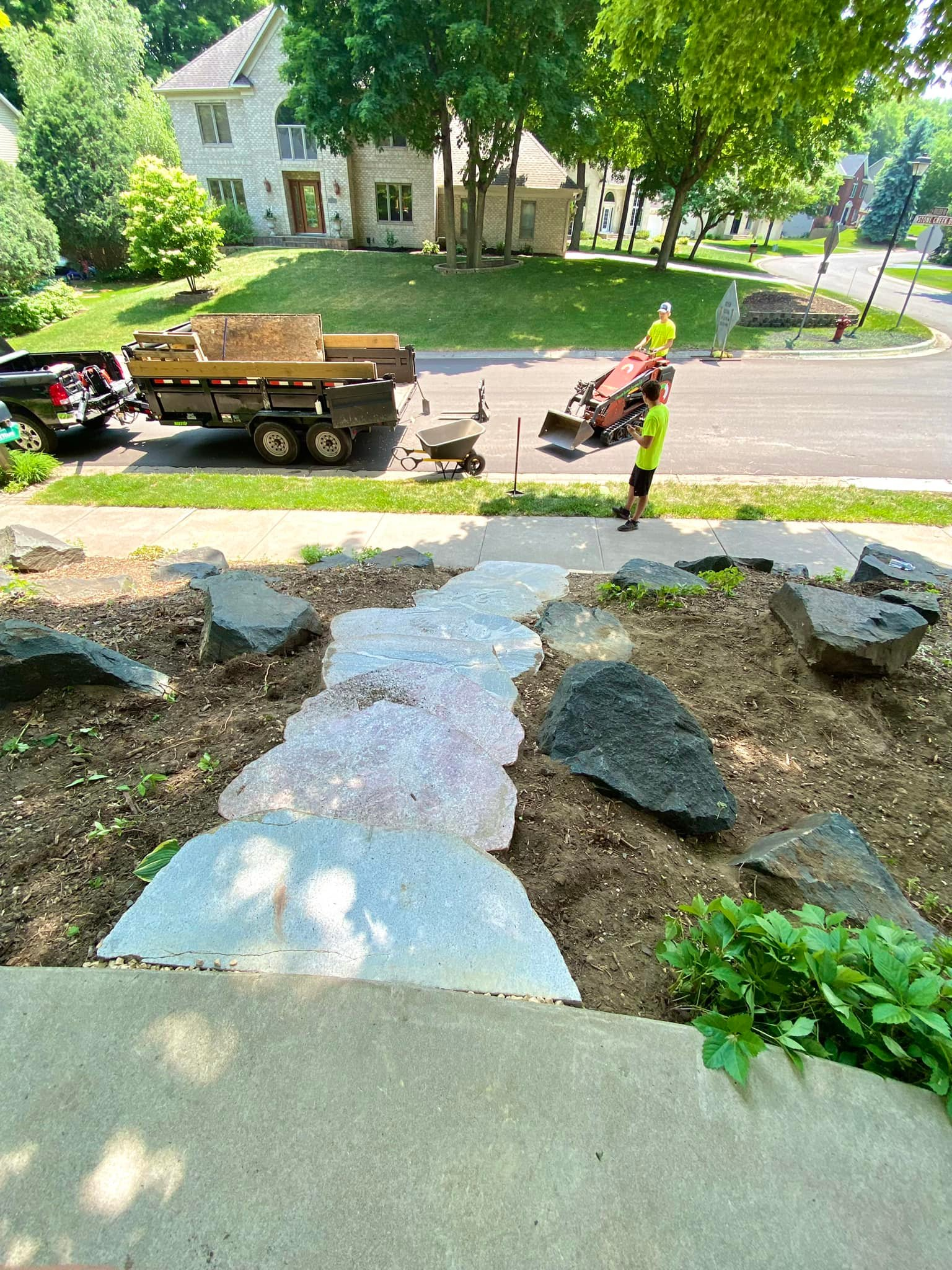 Workers installing a stone pathway in a yard. A truck with a trailer and a wheelbarrow are nearby.