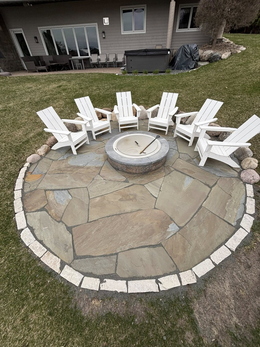 Round stone fire pit with white Adirondack chairs on a stone patio in a backyard.