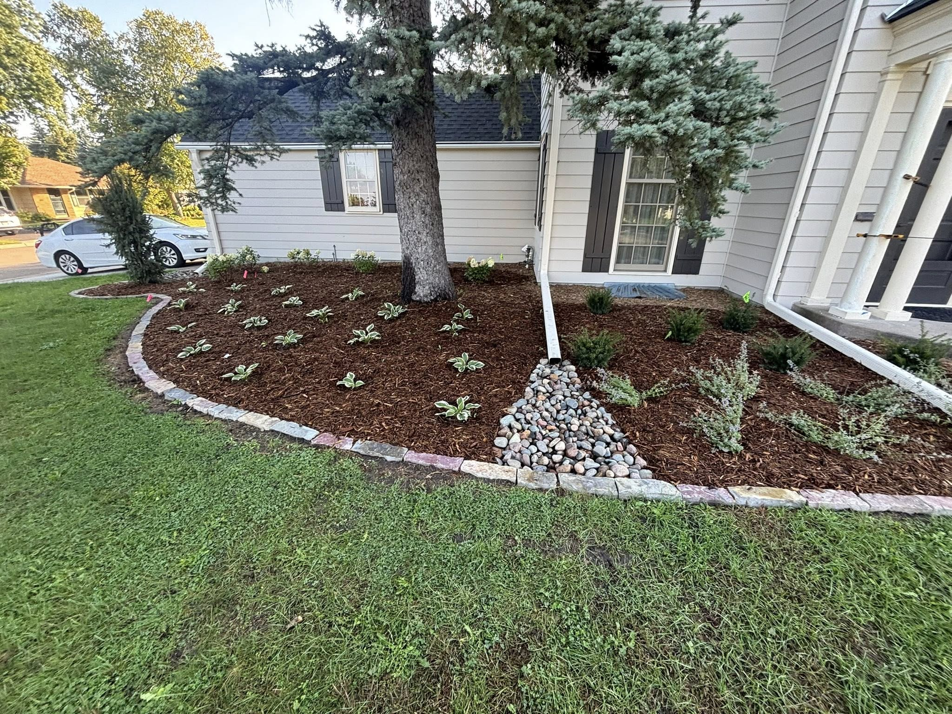 A house with a landscaped flowerbed. Brown mulch, small plants, and a tree trunk border the house's exterior.