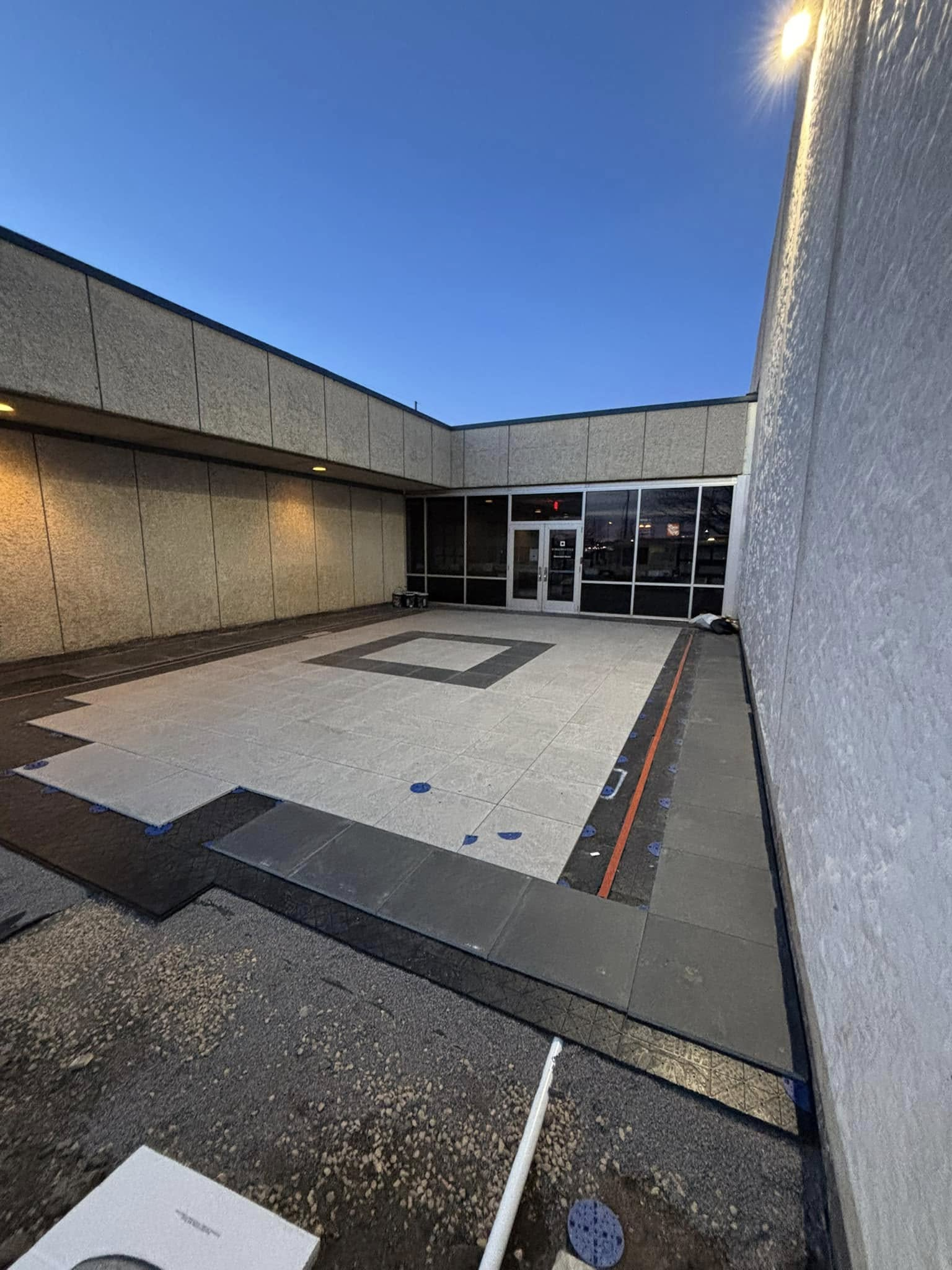 Outdoor plaza with construction. Concrete floor, gray tiles, entrance with glass doors, and a clear, blue sky.