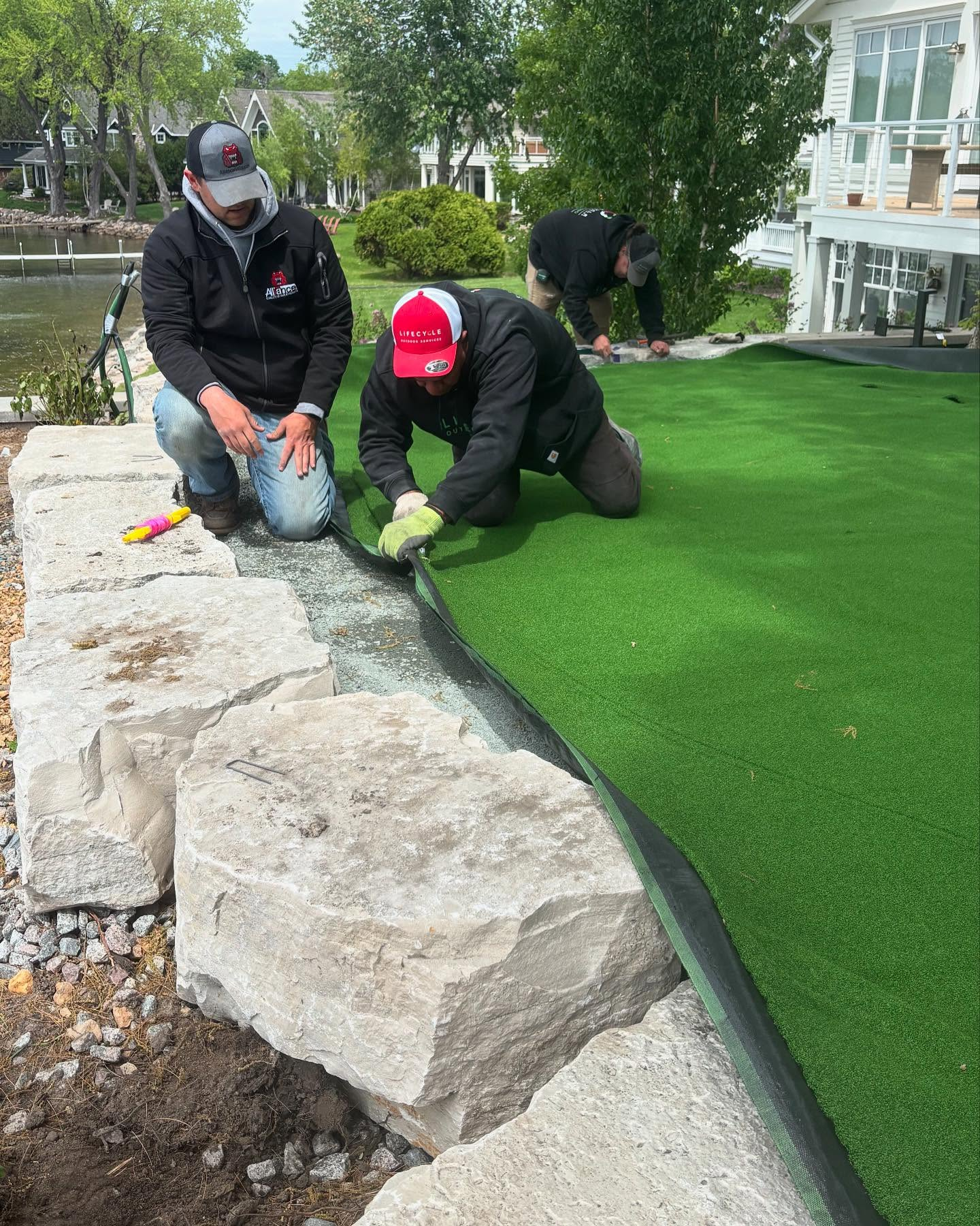 Three people installing artificial turf by a stone wall near a lake; one kneels, two bend over the material.