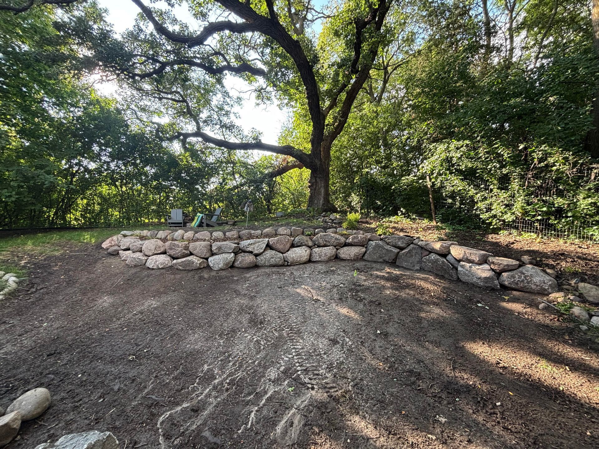 A tiered stone wall encircles a freshly tilled garden bed under a large tree in a wooded area.