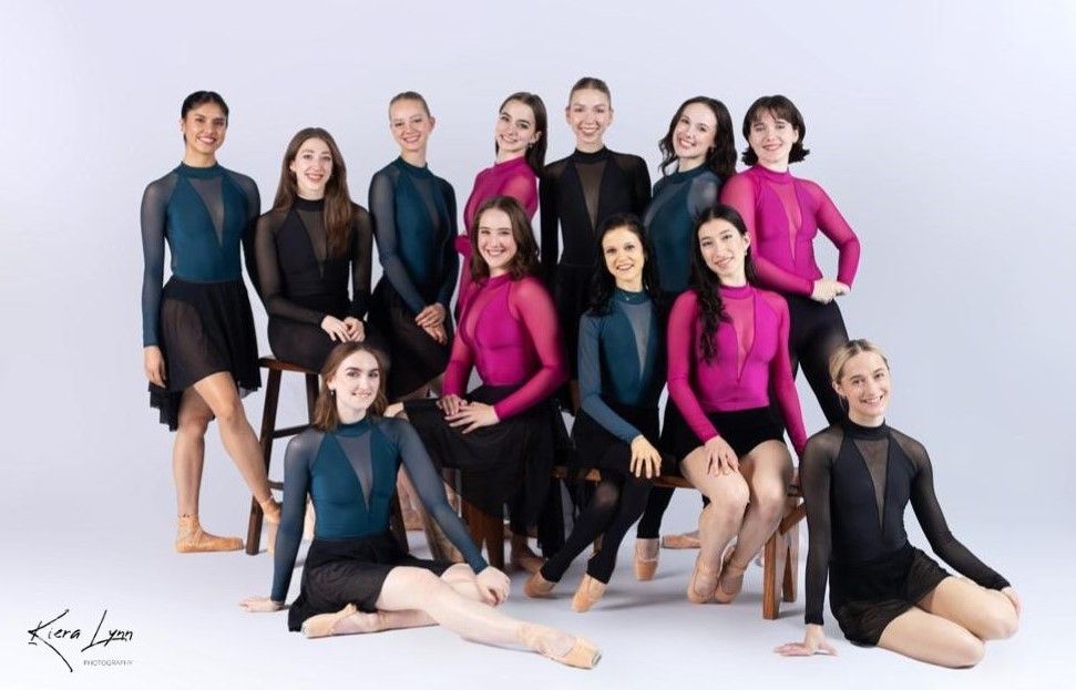 Group of dancers in various poses, wearing teal and pink long-sleeved leotards with black skirts and shorts.