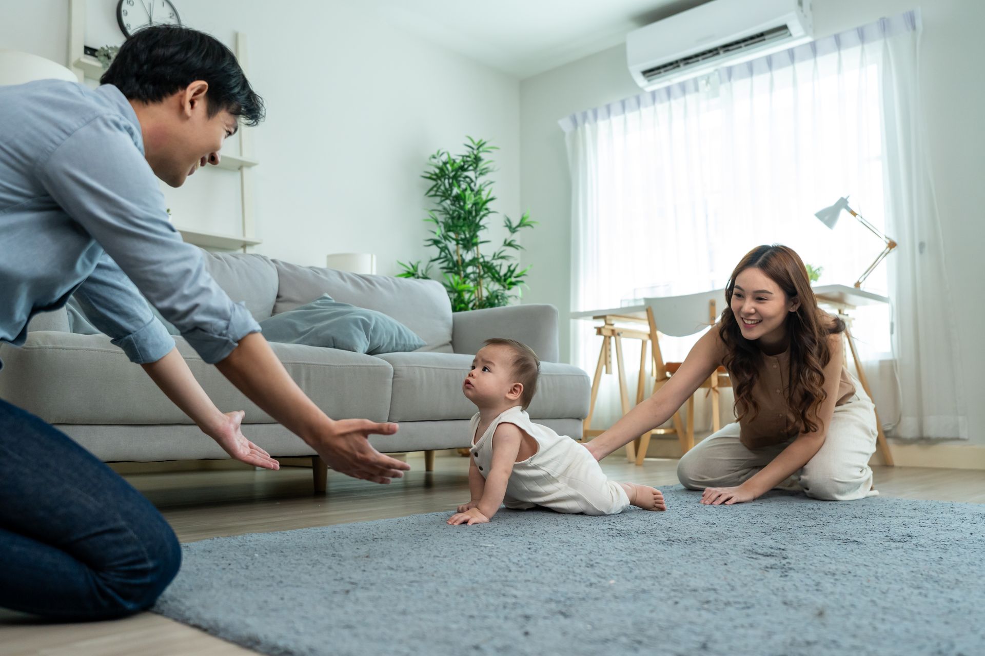 Family on a carpeted floor in a bright living room, helping a baby crawl toward them.
