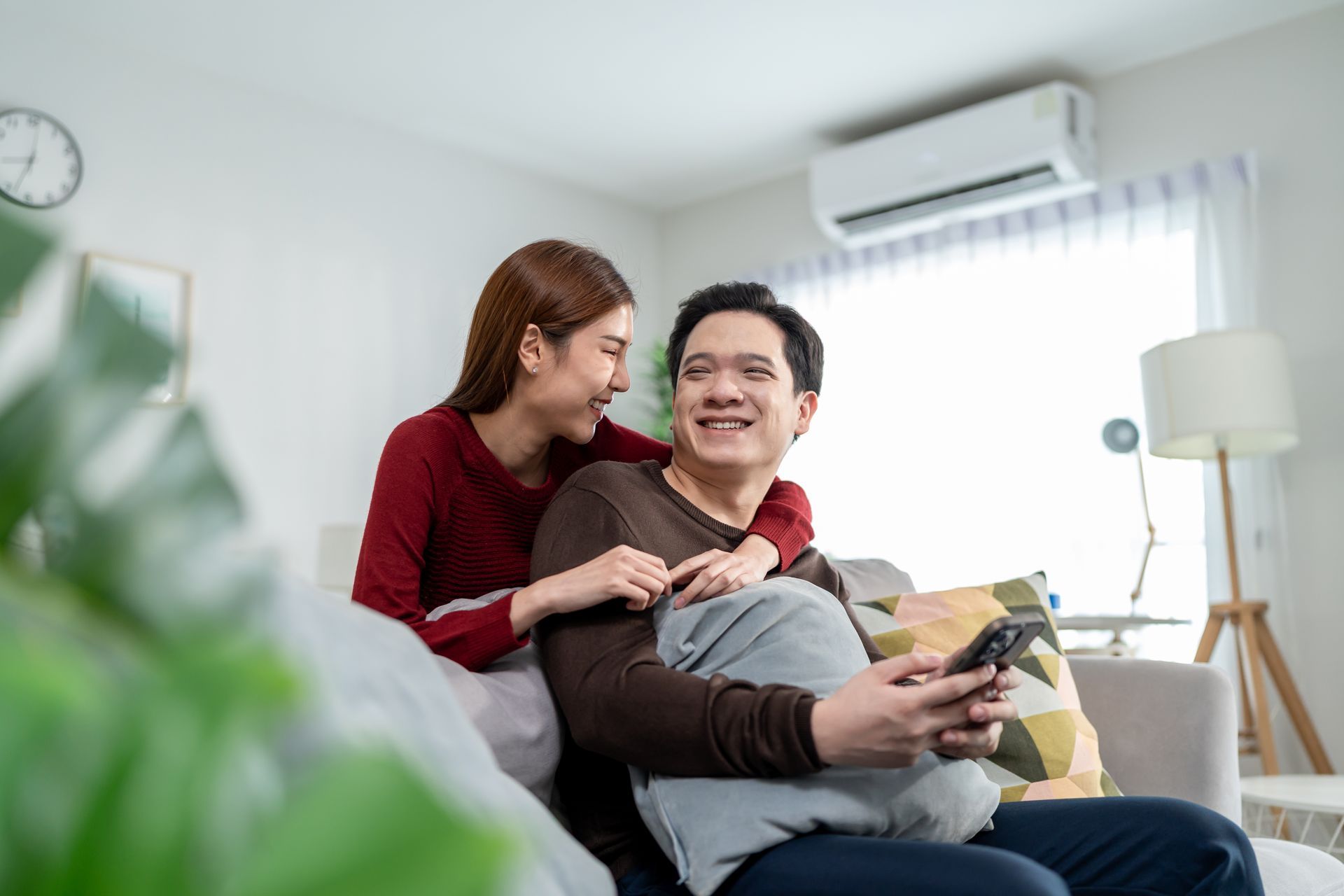 Two people smiling on a couch in a bright living room, one hugging the other with a tablet on his lap.