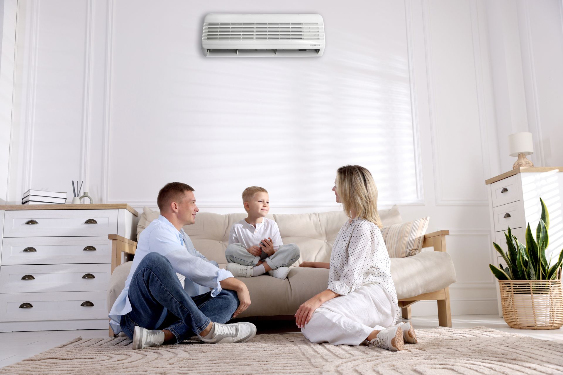 Three people sitting on the floor in a bright living room near a beige sofa and wall-mounted air conditioner