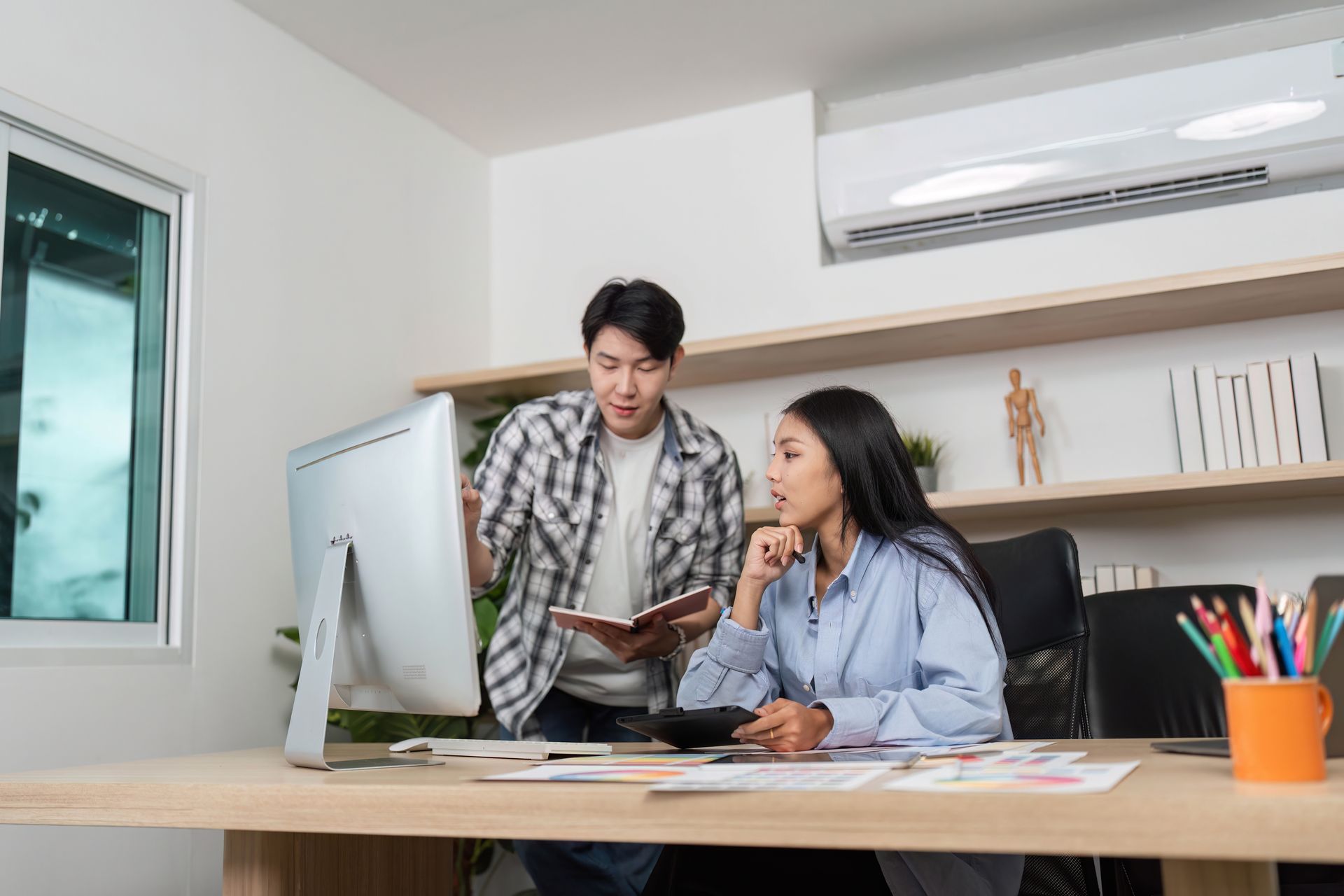 Two coworkers reviewing documents at a desk in a bright office with a computer and air conditioner.