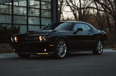 A sleek, black Dodge Challenger parked on a paved surface near a glass building with trees in the background.