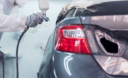 Person in protective suit spray painting the rear of a gray car in a workshop.
