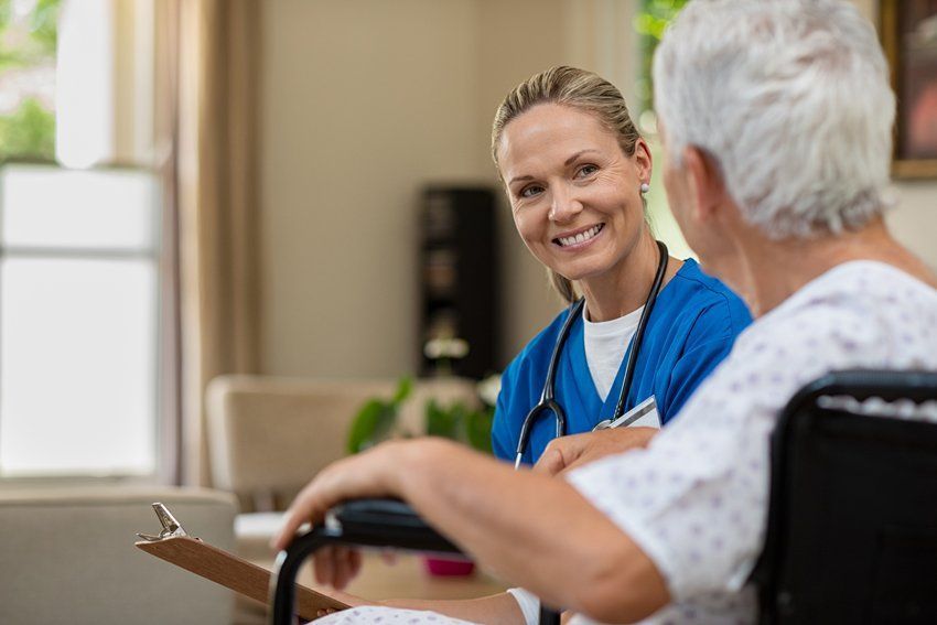 Nurse in blue scrubs smiles at a patient in a wheelchair, holding a clipboard in a living room.