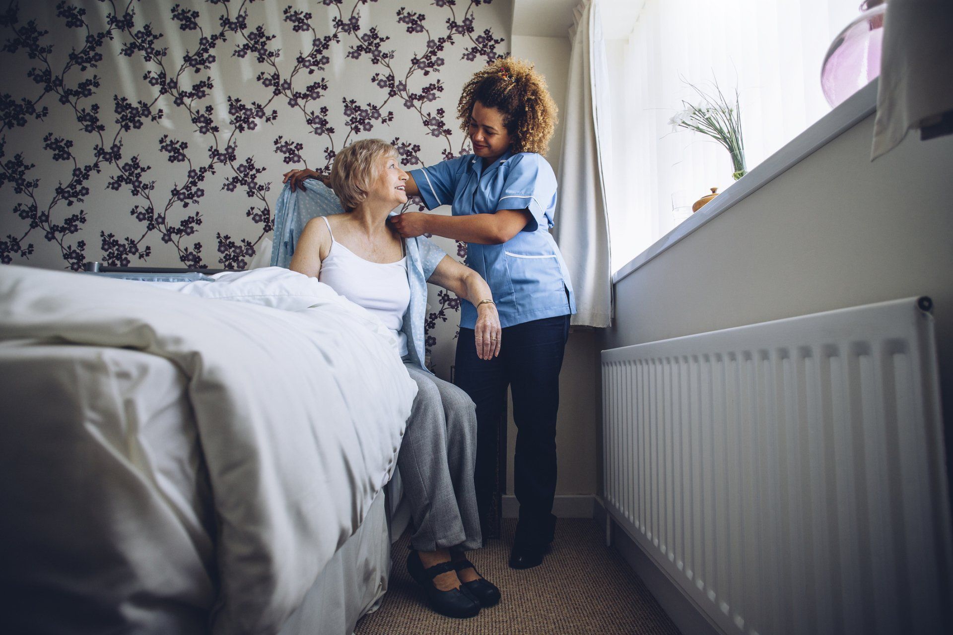 Nurse assisting a person sitting on a bed, adjusting their clothing near a window in a bedroom. Nurse assisting a person sitting on a bed, adjusting their clothing near a window in a bedroom.