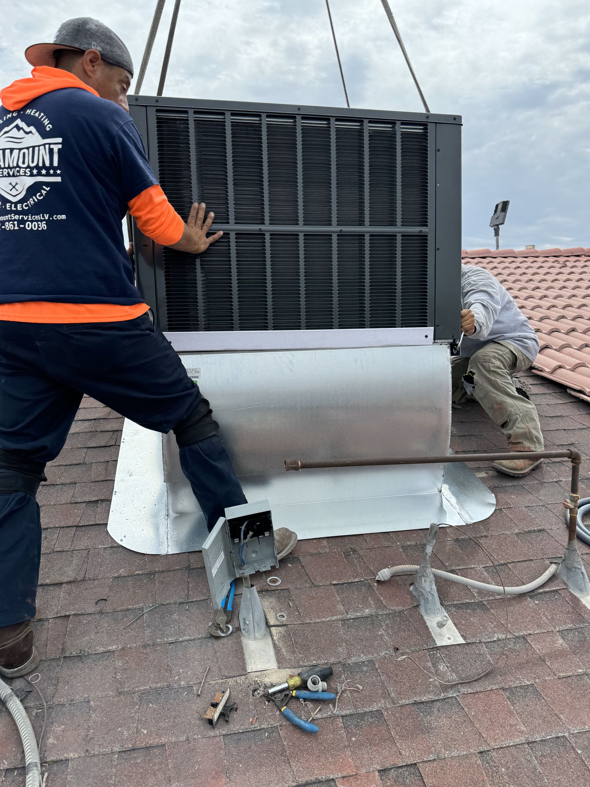 Two men install an air conditioning unit on a rooftop. One man guides it while the other preps the area, all under a cloudy sky.