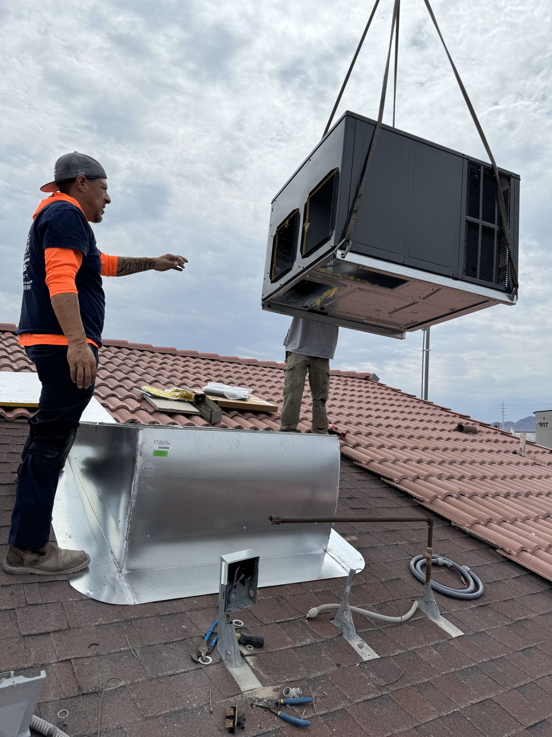 Two workers install an HVAC unit on a rooftop. A man points, guiding the unit lowered by a crane. The setting is outdoors.
