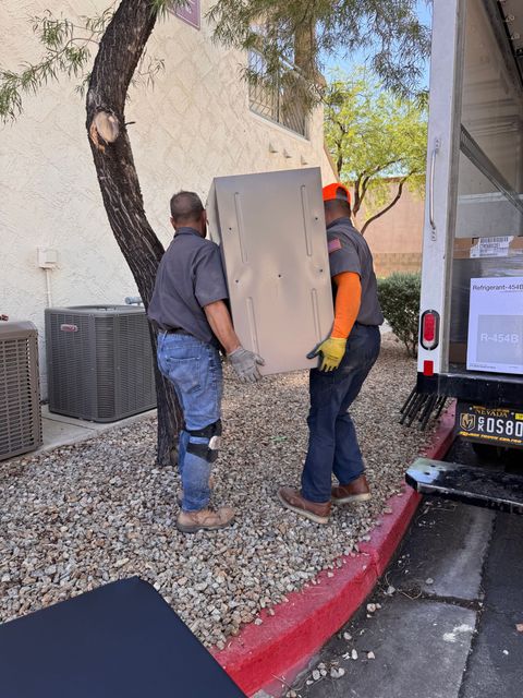 Two workers in uniforms carry a large boxed appliance from a delivery truck parked on a gravel lot near a building.